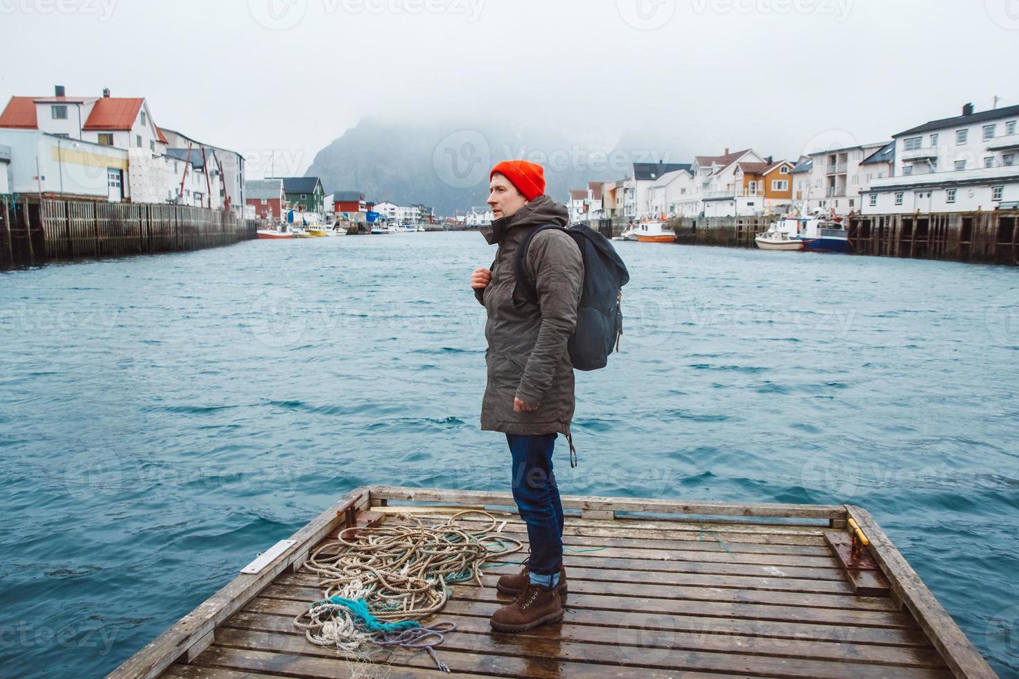 Reisender Mann mit einem Rucksack, der auf Woode Pier auf dem Hintergrund der Fischerhäuser Berg und See steht foto