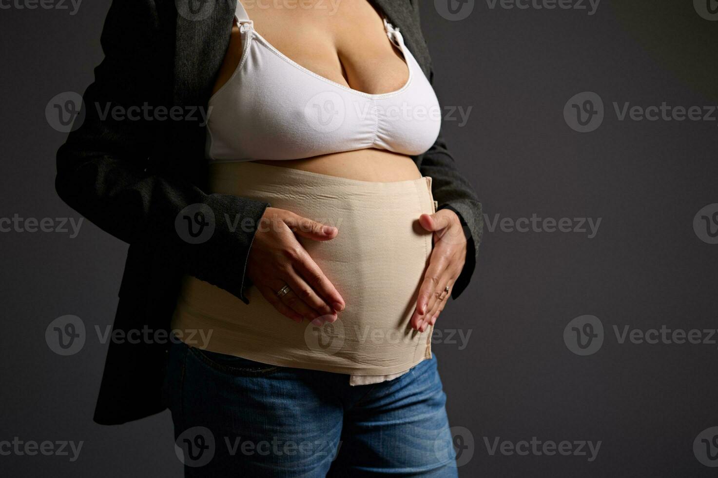 abgeschnitten Aussicht von ein jung Frau tragen elastisch Binde auf ihr postnatal Bauch, lächelnd suchen beim Kamera, grau Hintergrund foto