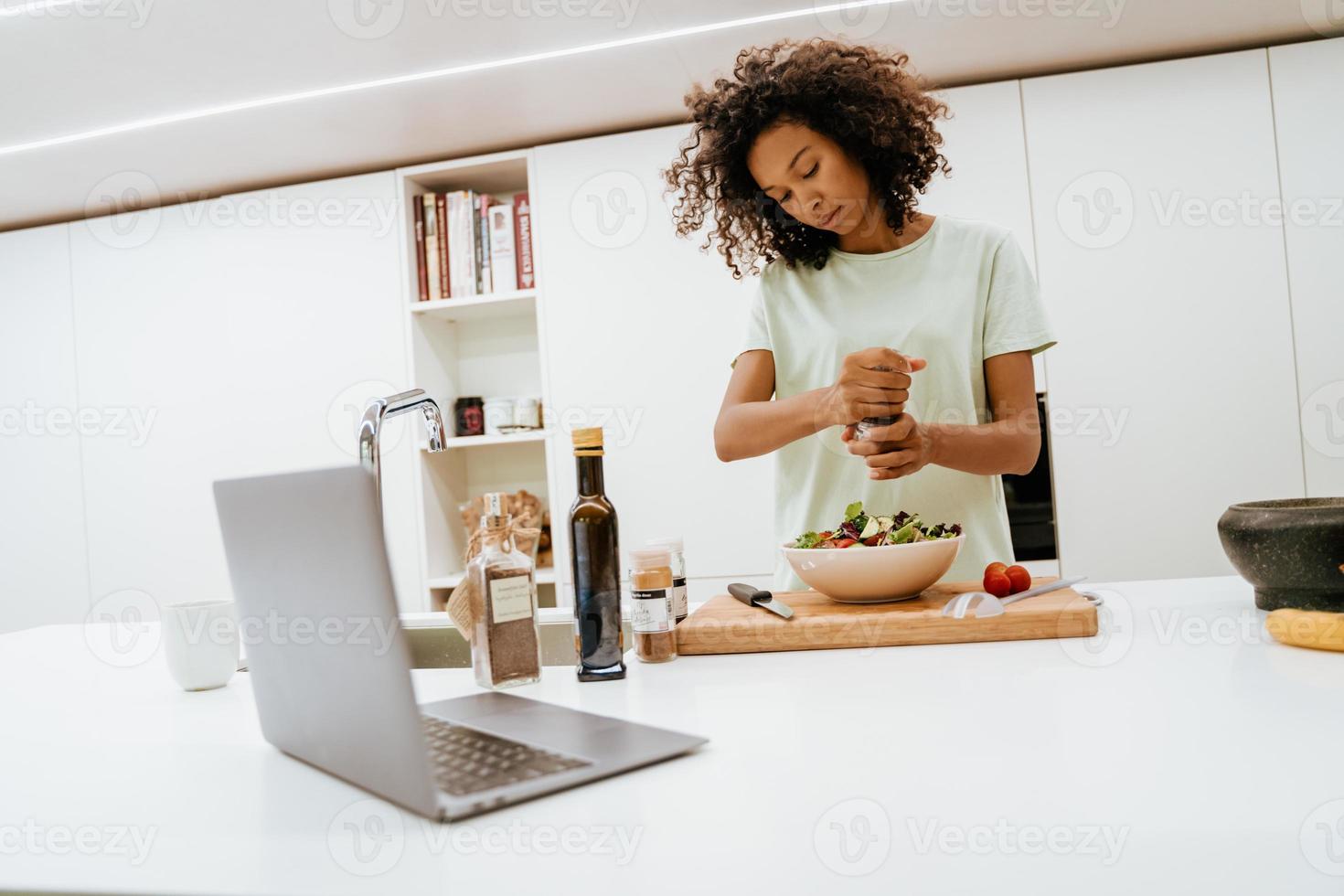 Schwarze junge Frau macht Salat, während sie Laptop in der Küche benutzt foto