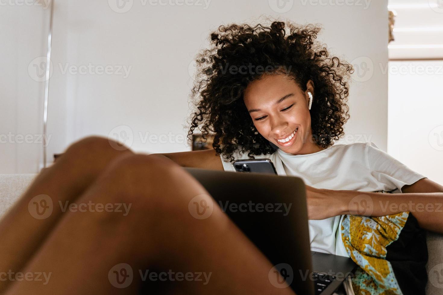 Schwarze junge Frau mit Handy und Laptop beim Ausruhen auf dem Sofa foto