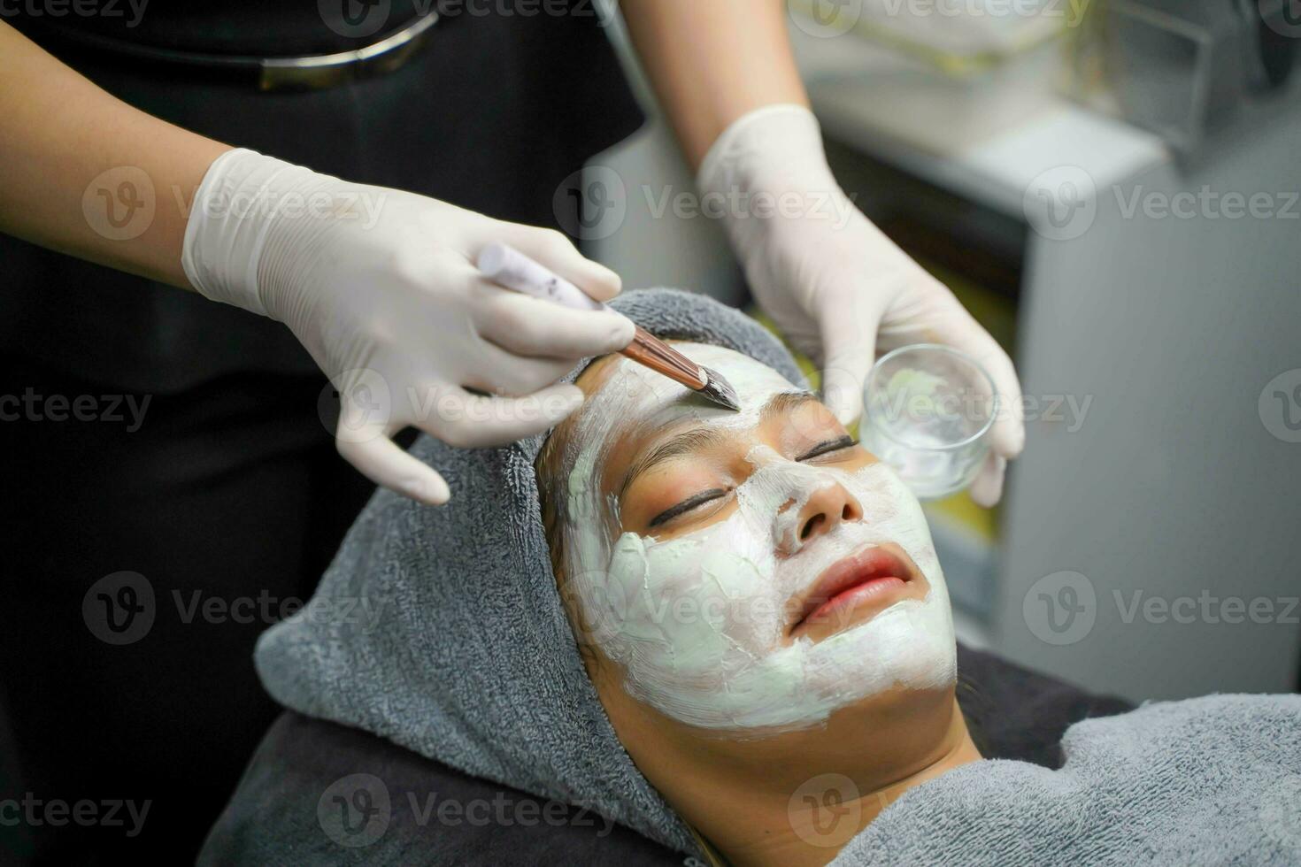 Nahansicht Kosmetikerin ist bewirbt sich Gesichts- Weiß Maske Sahne auf Frau Klient Gesicht im Schönheit Klinik. foto