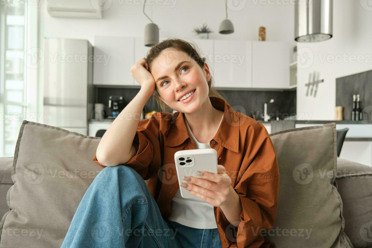 lächelnd Brünette Frau Sitzung auf Sofa mit Smartphone, suchen nachdenklich, Denken während mit Handy, Mobiltelefon Telefon Anwendung, Bestellung auf Anwendung, tun online Einkaufen und ruhen beim Zuhause foto