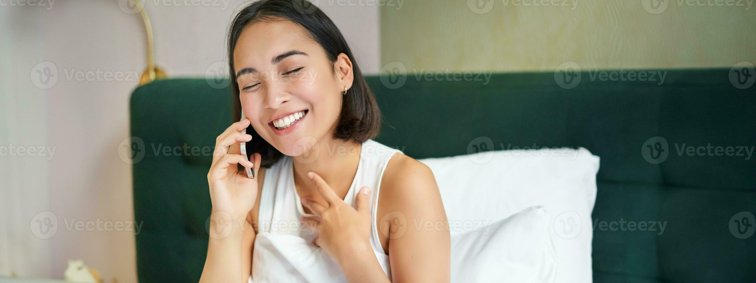 schließen oben Porträt von süß asiatisch Mädchen im Bett, reden auf Handy, Mobiltelefon Telefon mit glücklich lächelnd Gesicht. Frau aufwachen oben und Herstellung ein Telefon Anruf foto