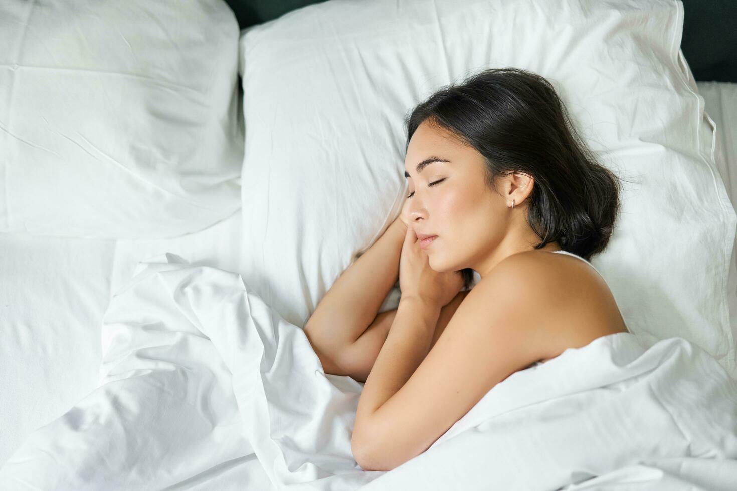 oben Aussicht Bild von asiatisch Frau Schlafen allein im König Größe Bett auf Weiß Kissen. jung Mädchen Lügen im ihr Schlafzimmer mit Augen geschlossen, Morgen Sonnenlicht scheint im Zimmer foto