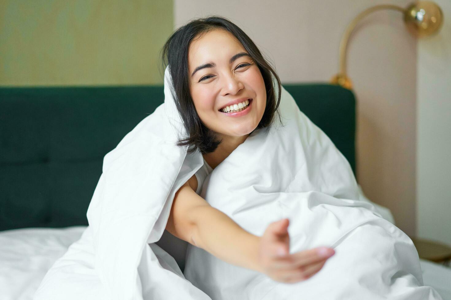 schön asiatisch Frau Sitzung auf Bett, bedeckt mit Weiß Bettdecke, lächelnd, genießen glücklich Wochenende Morgen, Lachen beim Kamera foto