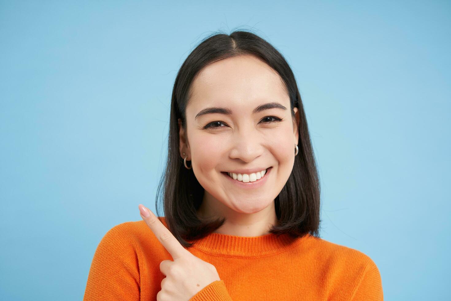 schließen oben Schuss von lächelnd asiatisch Mädchen, Punkte Finger beim Oberer, höher links Ecke, zeigt an Banner, demonstriert Werbung, Blau Hintergrund foto