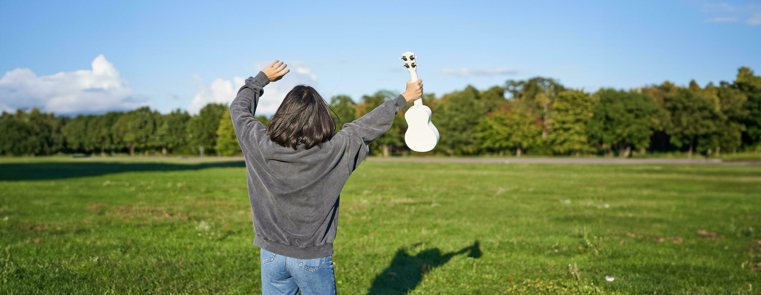 optimistisch jung Frau Tanzen mit ihr Musical Instrument. Mädchen erhöht ihr Ukulele oben und Pose im Park auf Grün Feld foto