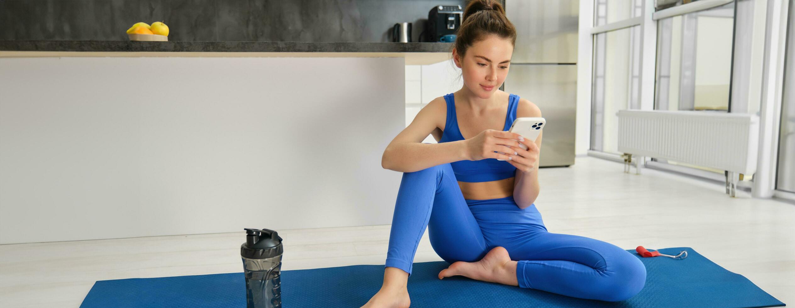 Porträt von jung Fitness Mädchen sitzt auf Yoga Matte mit Wasser Flasche, sieht aus beim Smartphone Anwendung, folgt Yoga Ausbildung Anleitung foto