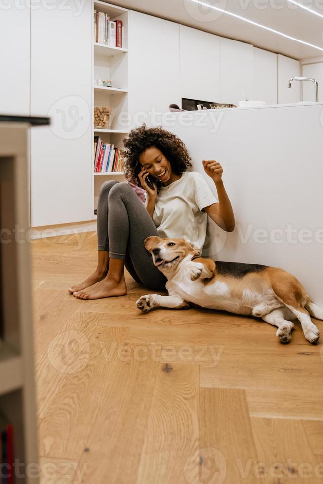 Schwarze junge Frau, die am Handy spricht und ihren Hund spielt foto