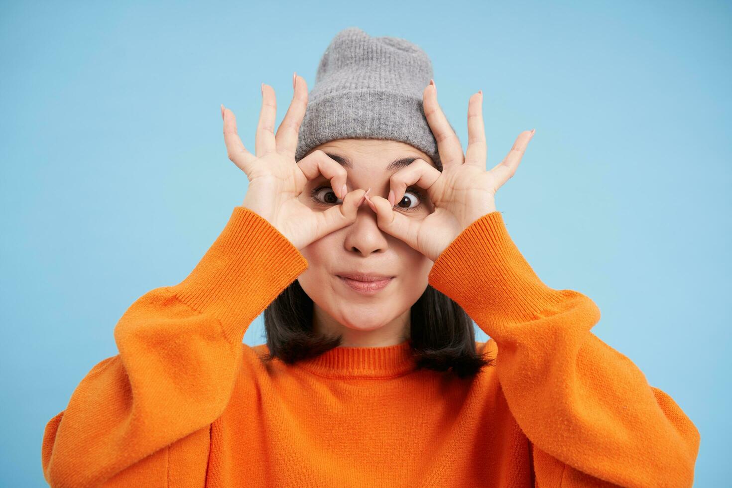 schließen oben Porträt von komisch Chinesisch Mädchen, sieht aus durch Hand Brille mit überrascht Gesicht Ausdruck, Stehen Über Blau Hintergrund foto