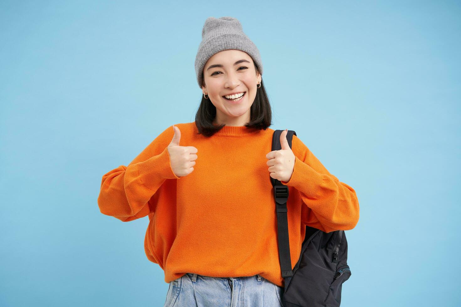 begeistert asiatisch Frau im Hut, hält Rucksack, zeigt an Daumen hoch, genehmigt etw, gibt positiv Rückmeldung, sagt Ja, Blau Hintergrund foto