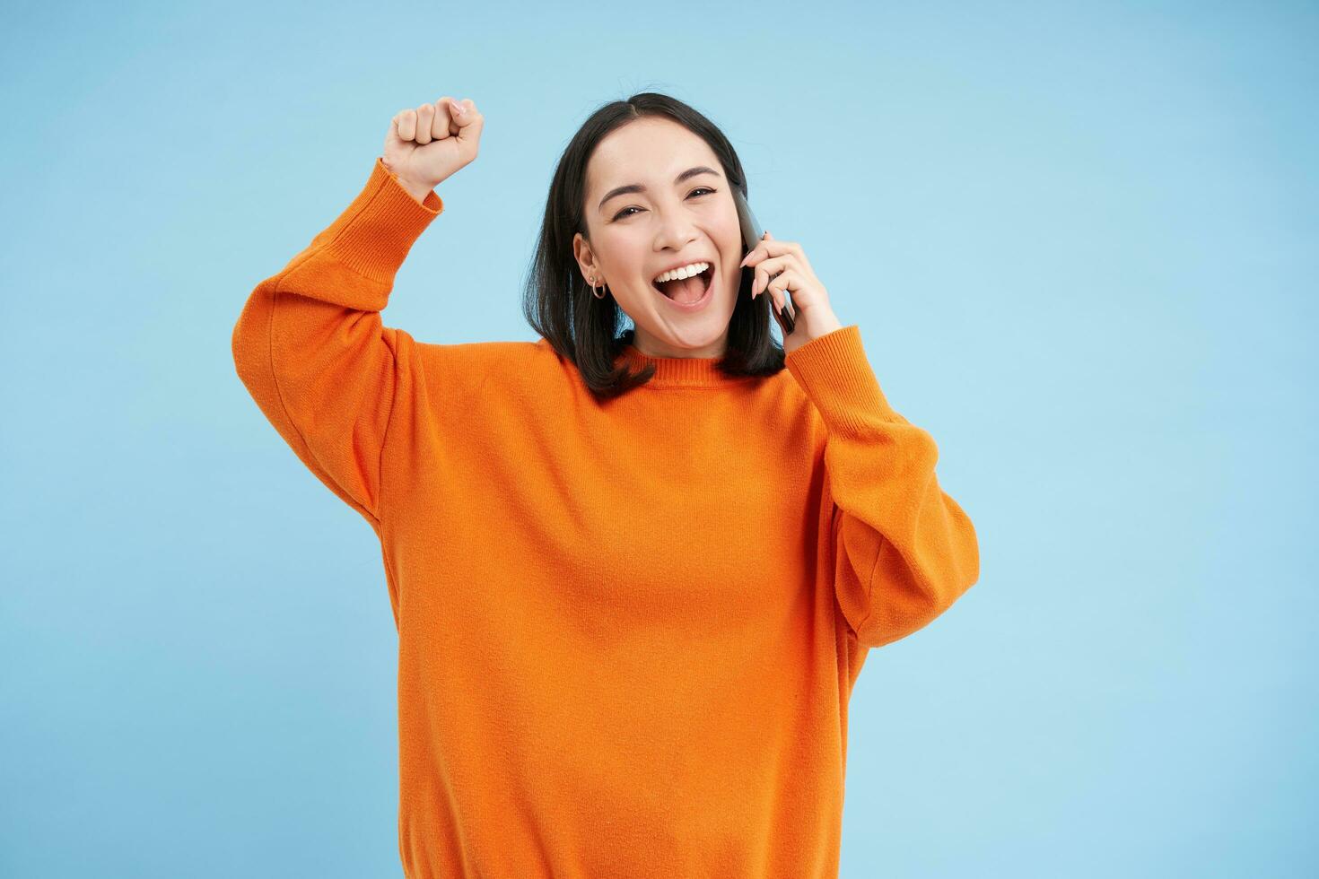 glücklich asiatisch Mädchen erhält Ausgezeichnet Nachrichten Über das Telefon, hält Handy, Mobiltelefon und tut Faust Pumpe, feiert Sieg, Leistung, Blau Studio Hintergrund foto