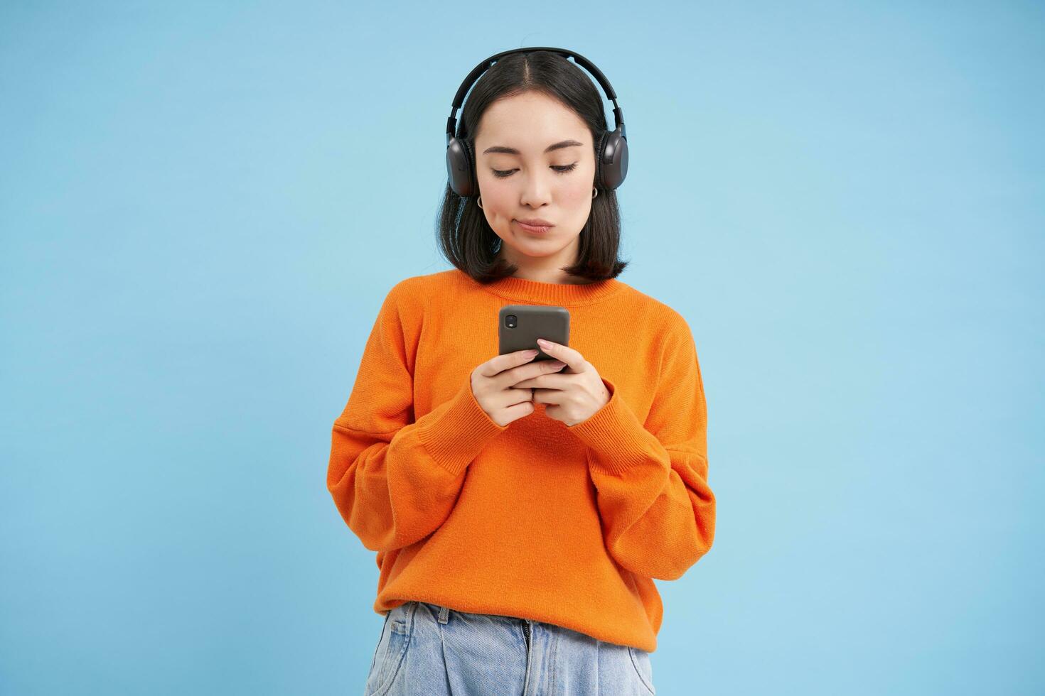 jung Frau im Kopfhörer und mit Smartphone hört zu Musik, genießt Singen Lieblings Lieder im ihr Wiedergabeliste, Blau Hintergrund foto