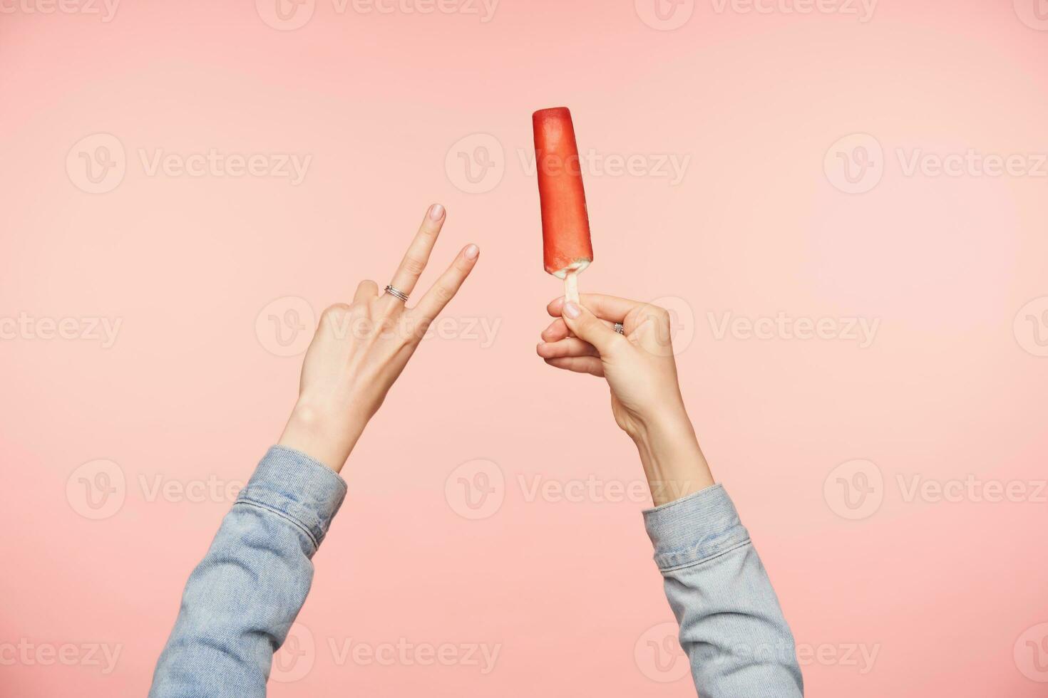 abgeschnitten Schuss von weiblich Hand Bildung Frieden Geste mit angehoben Hand und behalten rot Eis im andere Hand während Sein isoliert Über Rosa Hintergrund foto