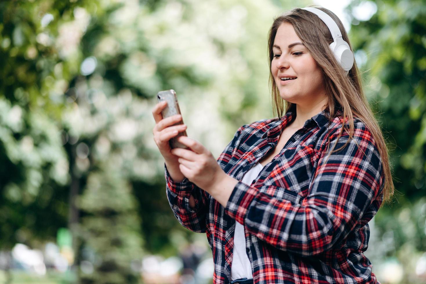 schön, Frau mit langen Haaren, in einem karierten Hemd, in Kopfhörern, mit einem Smartphone in der Hand im Freien foto