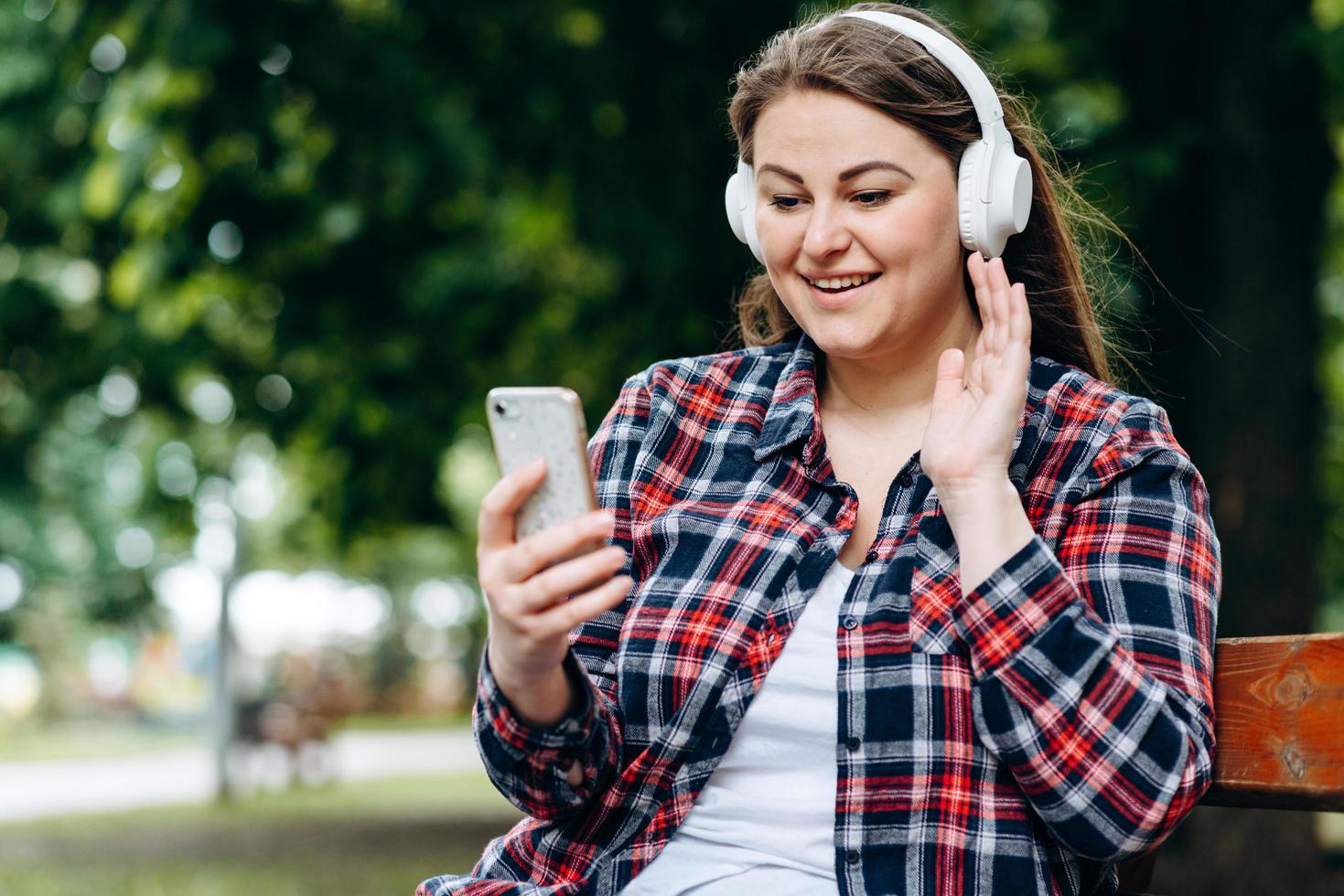 schöne, charmante Frau mit Ohrenschützern, die auf einer Bank sitzt, etwas über Kopfhörer hört und ein Smartphone hält foto