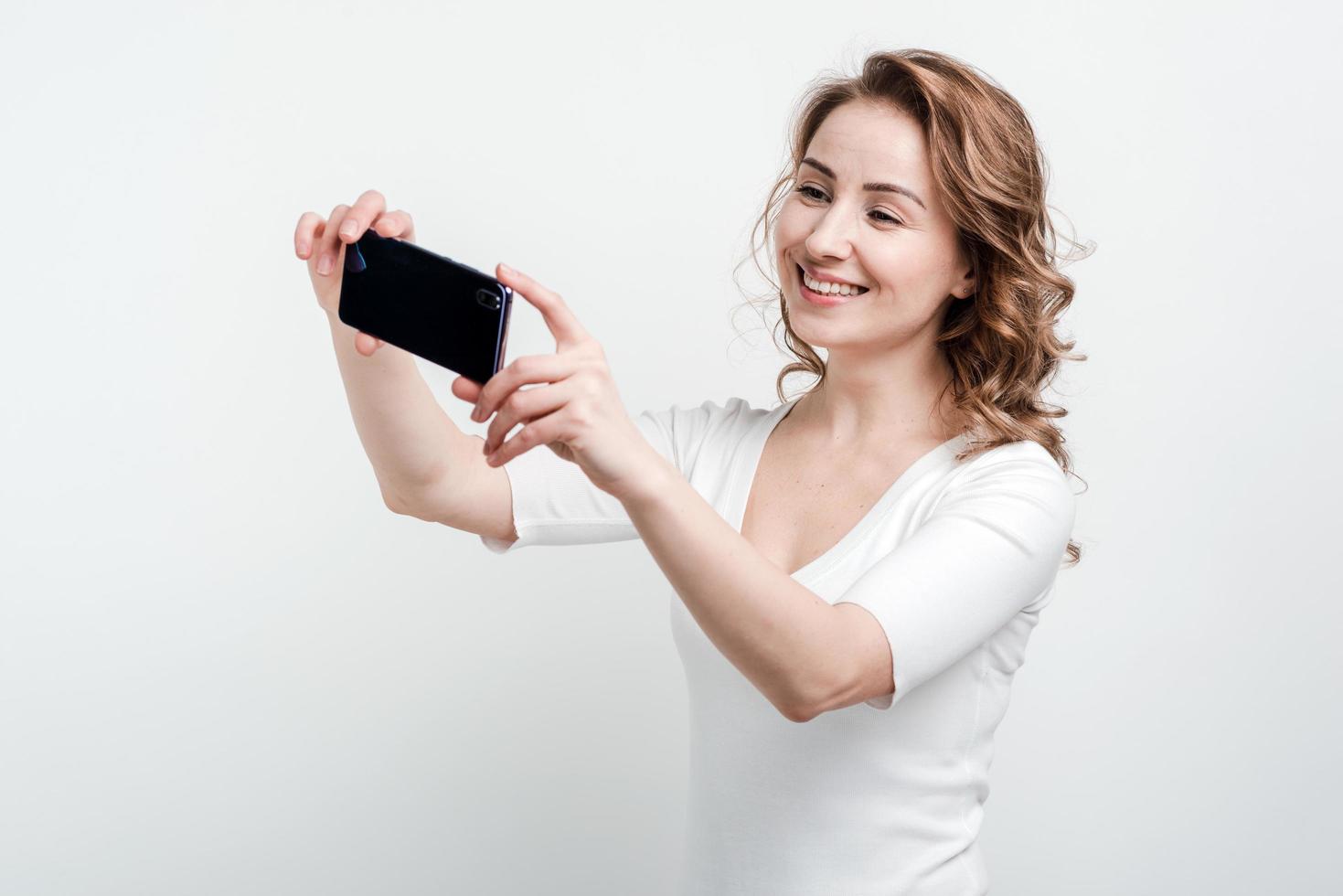 lächelndes Mädchen auf weißem Wandhintergrund macht Selfie am Telefon foto