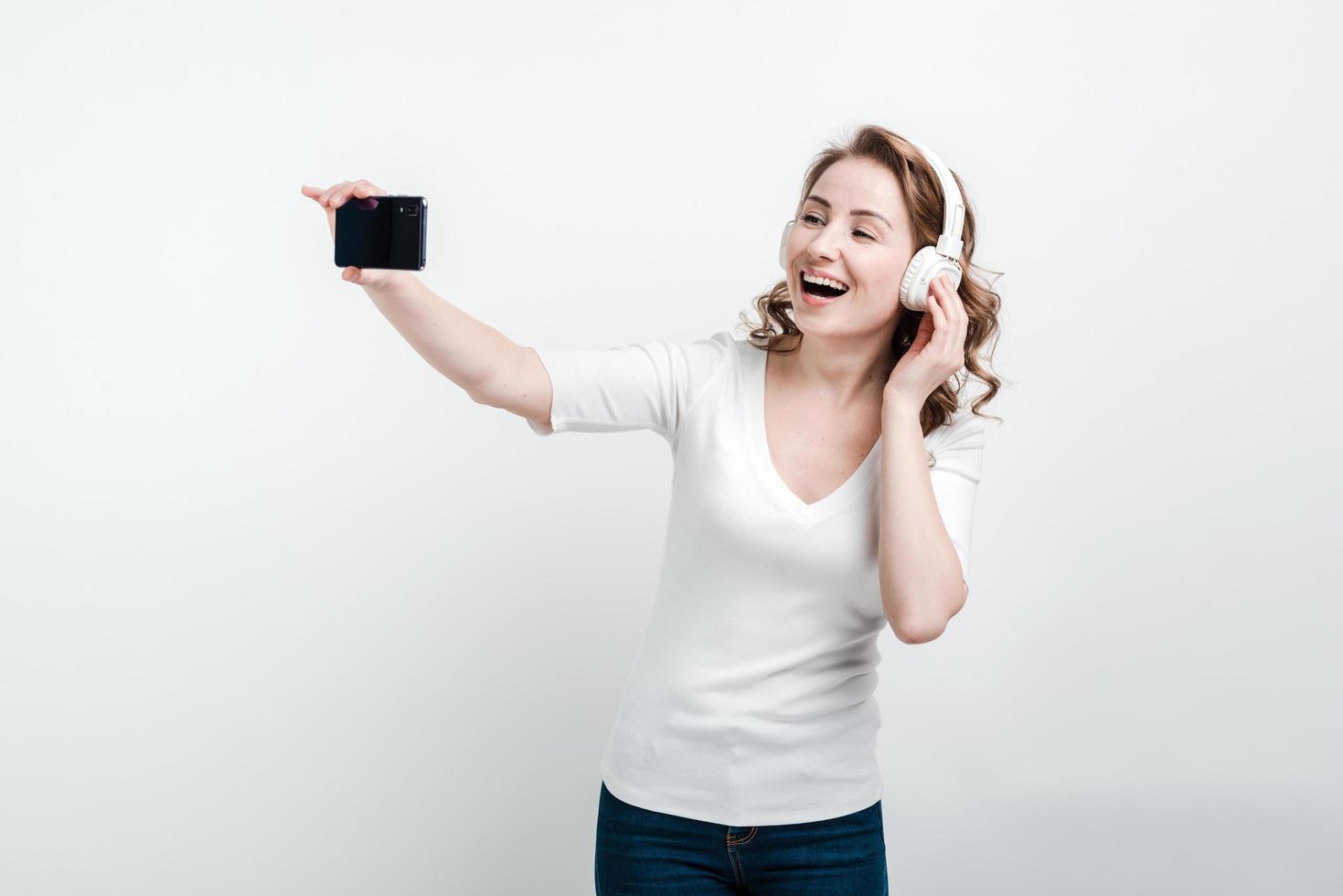 Mädchen mit weißen Kopfhörern lächelt und macht ein Selfie am Telefon foto