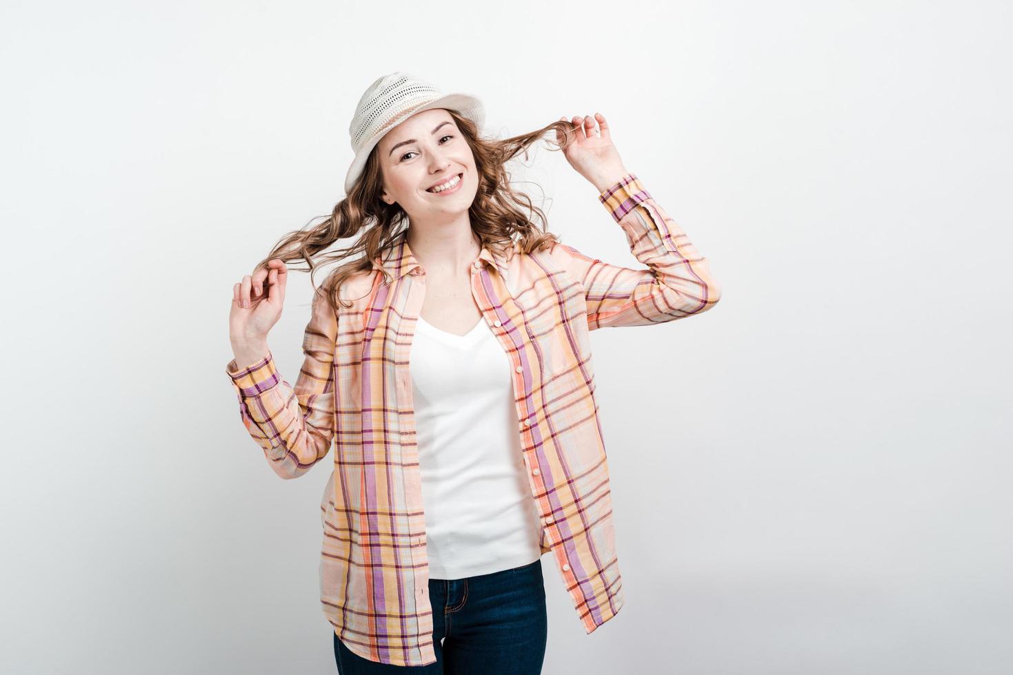 glückliche Frau mit Hut in Freizeitkleidung, die ihr Haar berührt, das über Studiohintergrund steht foto