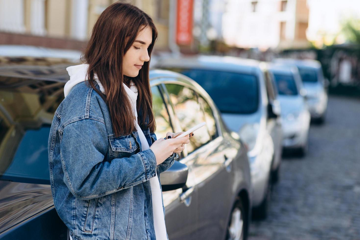 Mädchen auf einem städtischen Hintergrund, etwas am Telefon betrachtend, sich auf das Auto stützend foto
