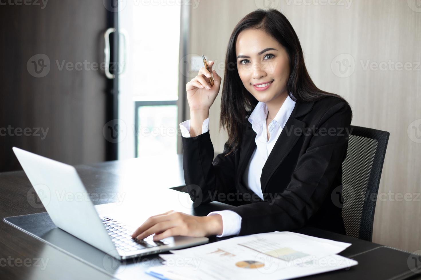 Asiatische Geschäftsfrauen, die einen Stift und Analysedokumente auf einem Bürotisch mit Laptop-Computer und grafischem Finanzdiagramm im Hintergrund halten foto