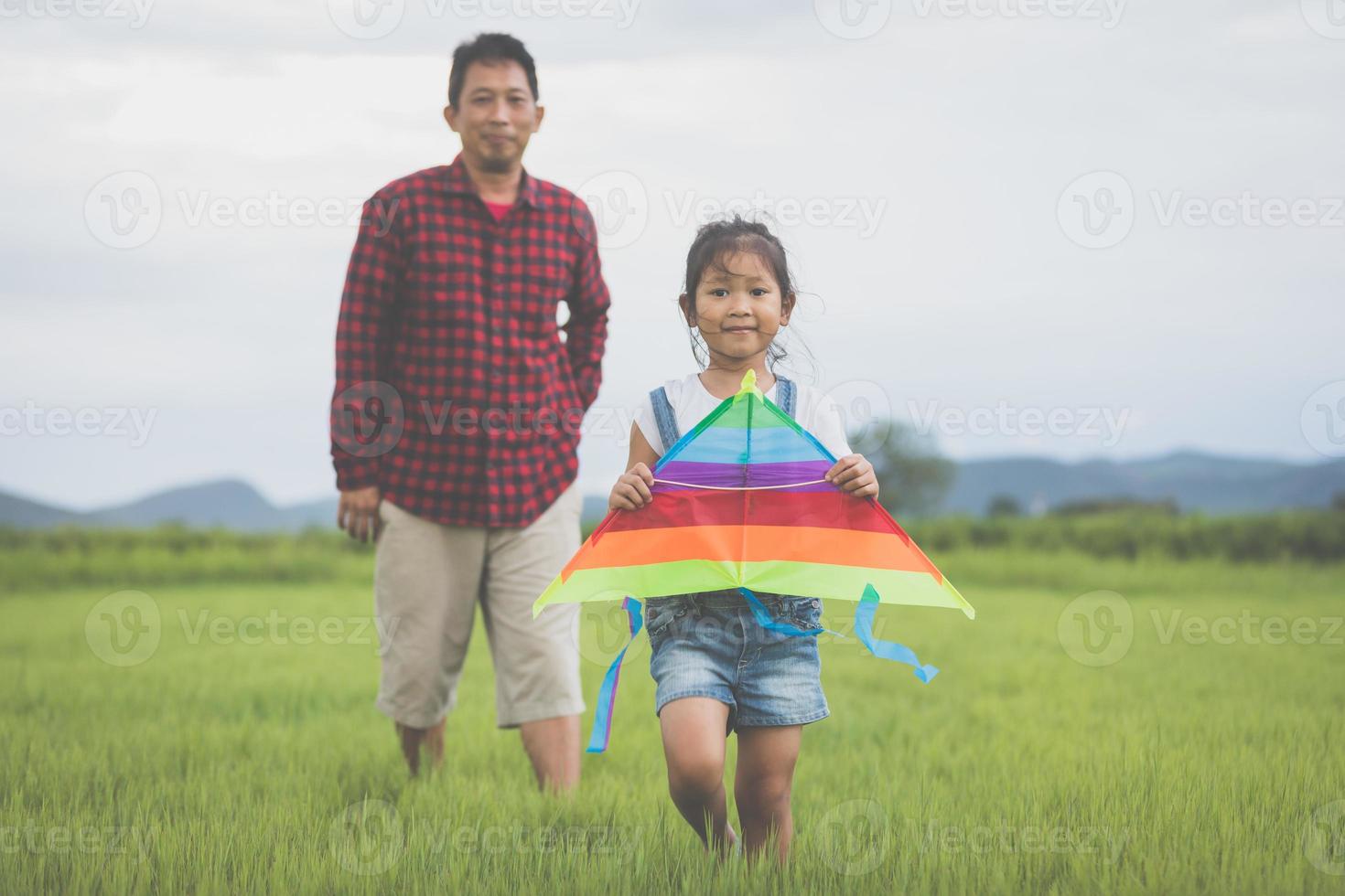 asiatisches kindermädchen und vater mit einem drachen, der im sommer in der natur auf der wiese läuft und glücklich ist foto