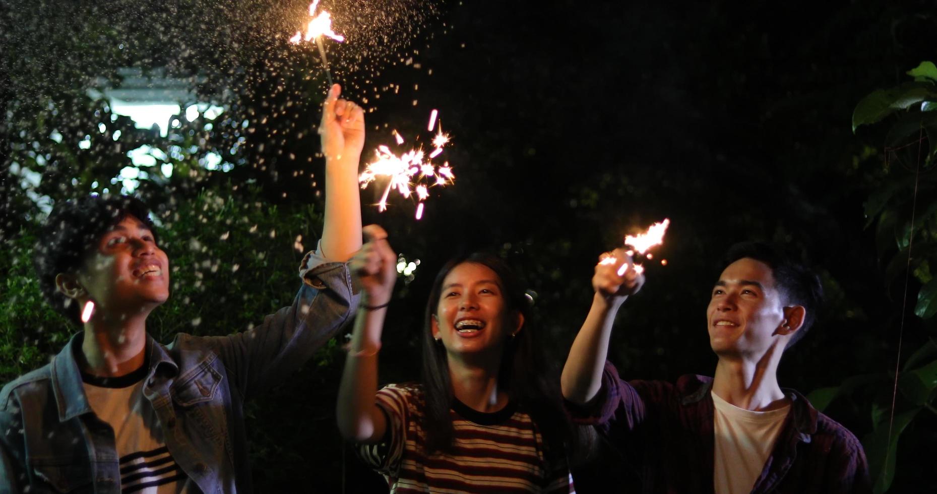 Asiatische Gruppe von Freunden beim Grillen im Freien, die mit alkoholischen Biergetränken lachen und eine Gruppe von Freunden zeigen, die sich nachts mit Wunderkerzen amüsieren, weicher Fokus foto