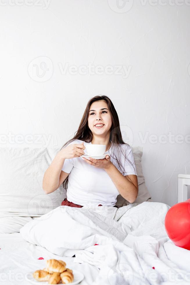 Junge brünette Frau sitzt wach im Bett mit roten herzförmigen Luftballons und Dekorationen, die Kaffee trinken und Croissants essen foto