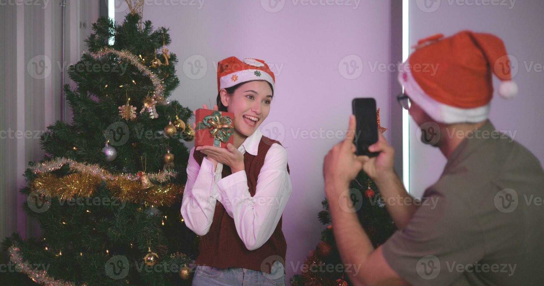 Porträt von jung asiatisch schön Paar nehmen Selfie und lächelnd während Sitzung mit dekoriert Weihnachten Baum im das Hintergrund, feiern Weihnachten und Neu Jahr. foto