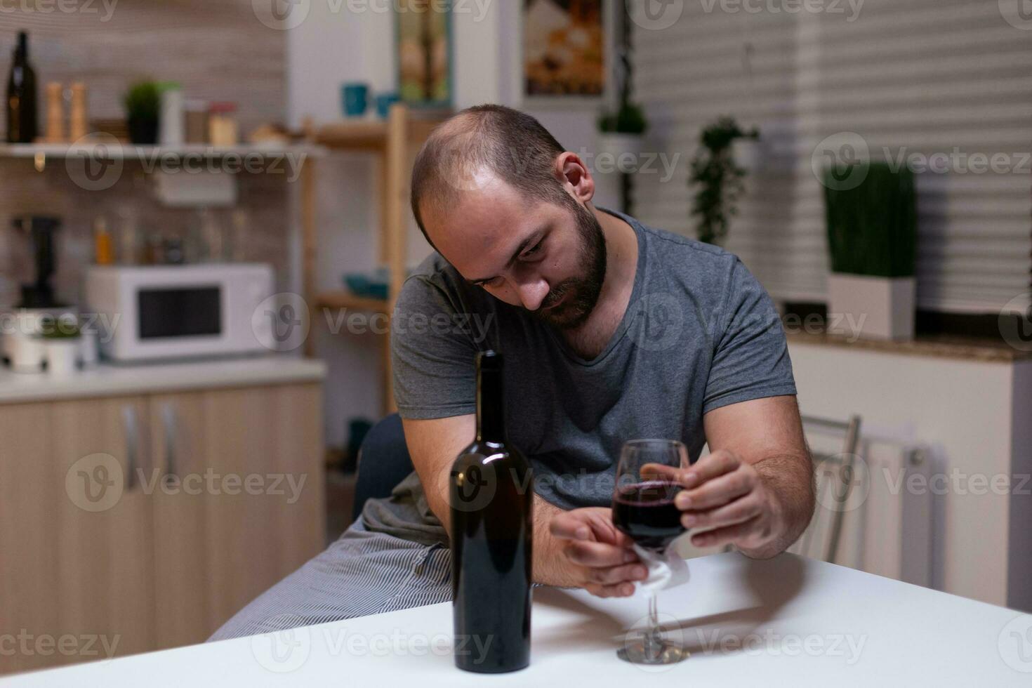 kaukasisch Mann halten Glas von Wein Sitzung im Küche beim heim. deprimiert Person Trinken Alkohol, Schnaps, alkoholisch Getränk allein während Gefühl berauscht mit Flasche von Alkohol foto