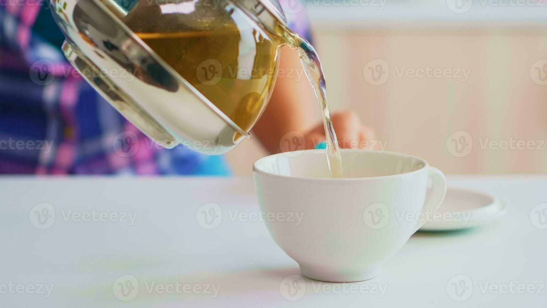 Grün Tee fließend von Teekanne im schleppend Bewegung. schließen oben von Tee von das Kessel langsam gießen in Porzellan Tasse im das Küche im das Morgen beim Frühstück, mit Teetasse und gesund Kräuter- Blätter. foto