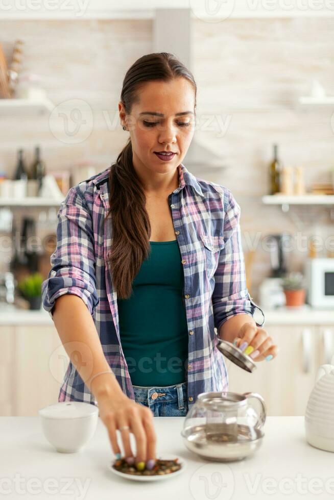 Frau mit aromatisch zum Vorbereitung von heiß Tee während Frühstück im heim. vorbereiten Tee im das Morgen, im ein modern Küche Sitzung in der Nähe von das Tisch. Putten mit Hände, gesund Kräuter- im Topf. foto