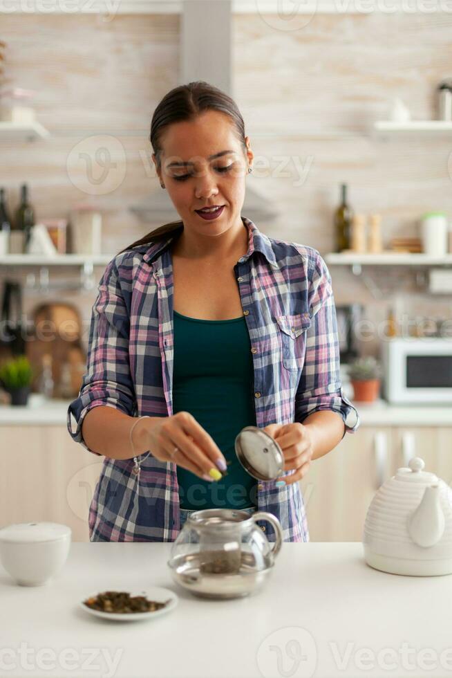 mit natürlich Kräuter im Küche zu bereiten Tee während Frühstück. vorbereiten Tee im das Morgen, im ein modern Küche Sitzung in der Nähe von das Tisch. Putten mit Hände, gesund Kräuter- im Topf. foto