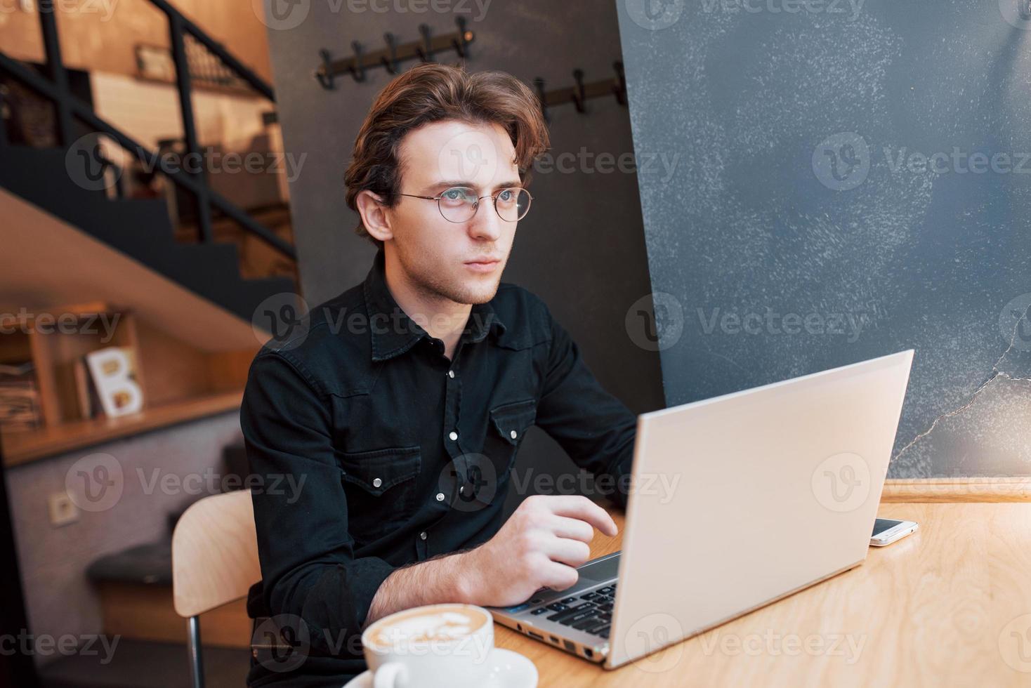 Kreativer Mann-Designer, der an seinem Laptop arbeitet, während er auf Bestellungen in seinem Lieblingscafé im Haus wartet, männlicher Student, der beim morgendlichen Frühstück im modernen Café-Interieur am Net-Book arbeitet foto