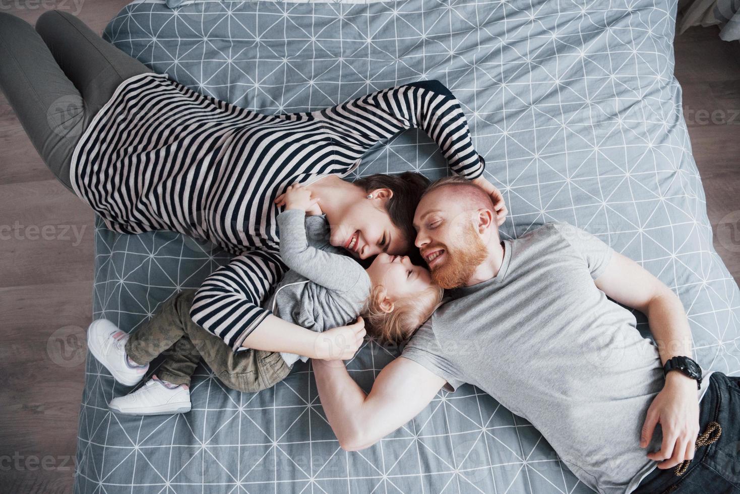 Blick von oben auf die schöne junge Mutter, den Vater und ihre Tochter, die in die Kamera schauen und lächeln, während sie Kopf an Kopf auf dem Bett liegen foto