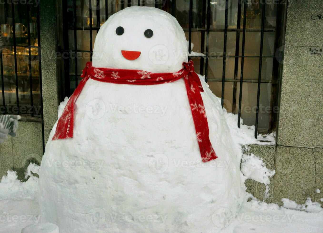 ein süß Schneemann tragen ein rot Schal auf Schnee Fußboden und Vorderseite von Zuhause Fenster im Winter Jahreszeit. foto