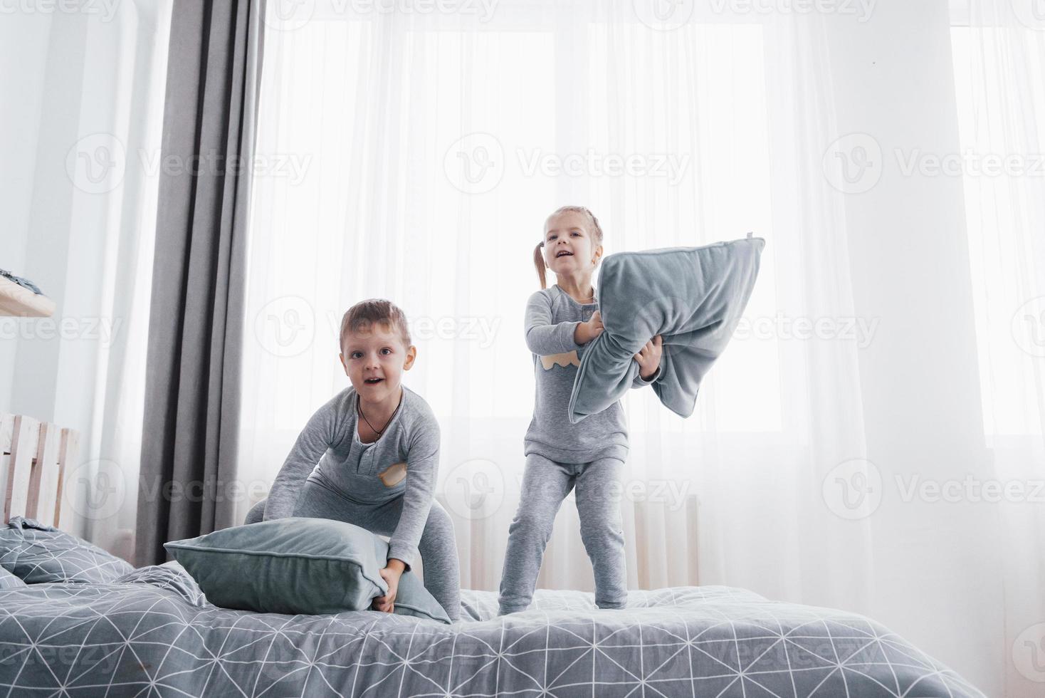 Kinder, die im Bett der Eltern spielen. Kinder wachen im sonnigen weißen Schlafzimmer auf. Junge und Mädchen spielen in passenden Pyjamas. Nachtwäsche und Bettzeug für Kind und Baby. Kinderzimmereinrichtung für Kleinkinder. Familienmorgen foto