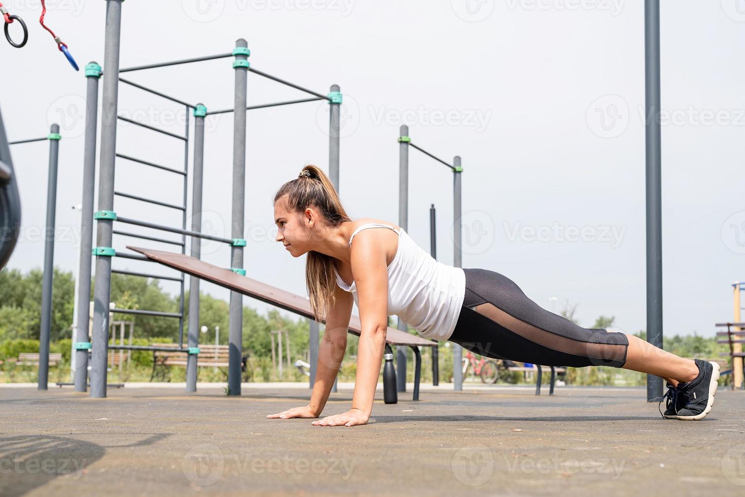 glückliche Frau, die an sonnigen Sommertagen auf dem Sportplatz trainiert und in Plankenposition steht foto