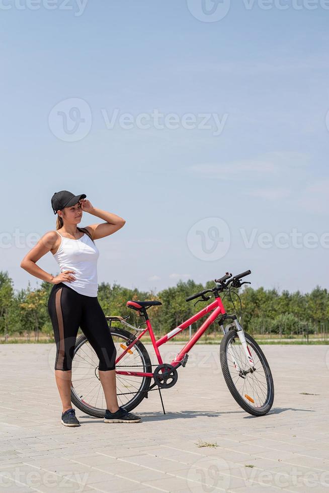 glückliche und müde Frau, die in einem Park an ihrem Fahrrad steht, sich ausruht, ihre Reise beendet und den Schweiß wischt foto