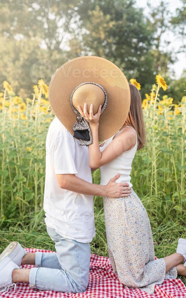 junges glückliches Paar, das sich auf einer Picknickdecke küsst und ihre Gesichter mit einem Sommerhut bedeckt foto