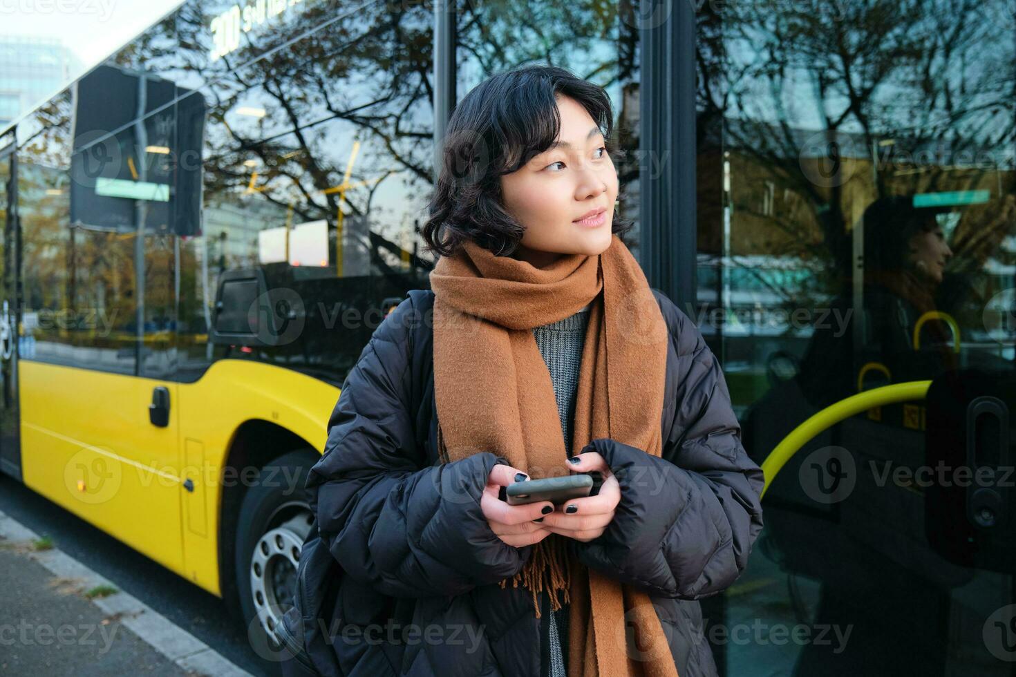 Porträt von Koreanisch Mädchen Kauf Fahrkarte zum Öffentlichkeit Transport online, mit Handy, Mobiltelefon Anwendung auf Bus stoppen, tragen Winter Kleider foto