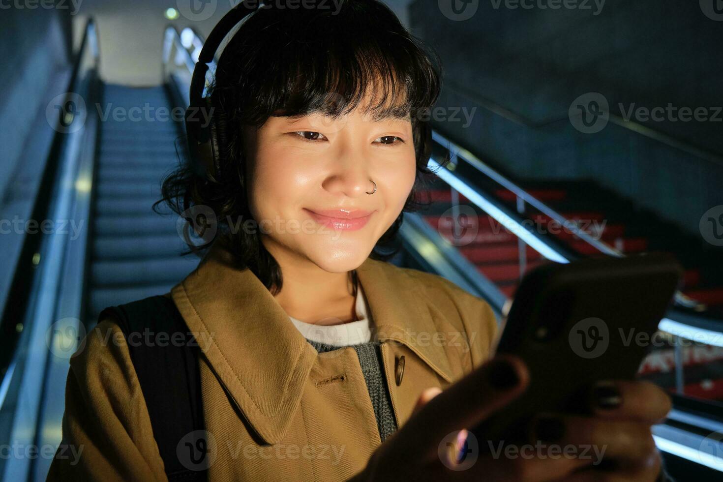 schließen oben von süß asiatisch Mädchen im Kopfhörer, Picks Lied zu Hör mal zu während pendeln, gehen Nieder Rolltreppe zu Metro, lächelnd wie lesen Botschaft auf Handy, Mobiltelefon Telefon foto