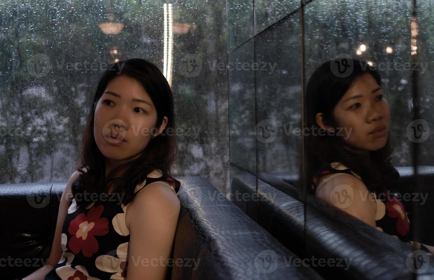 junge schöne asiatische frau mit traurigem und linigem gesicht, die am regnerischen tag im café mit reflexion vom spiegel sitzt foto