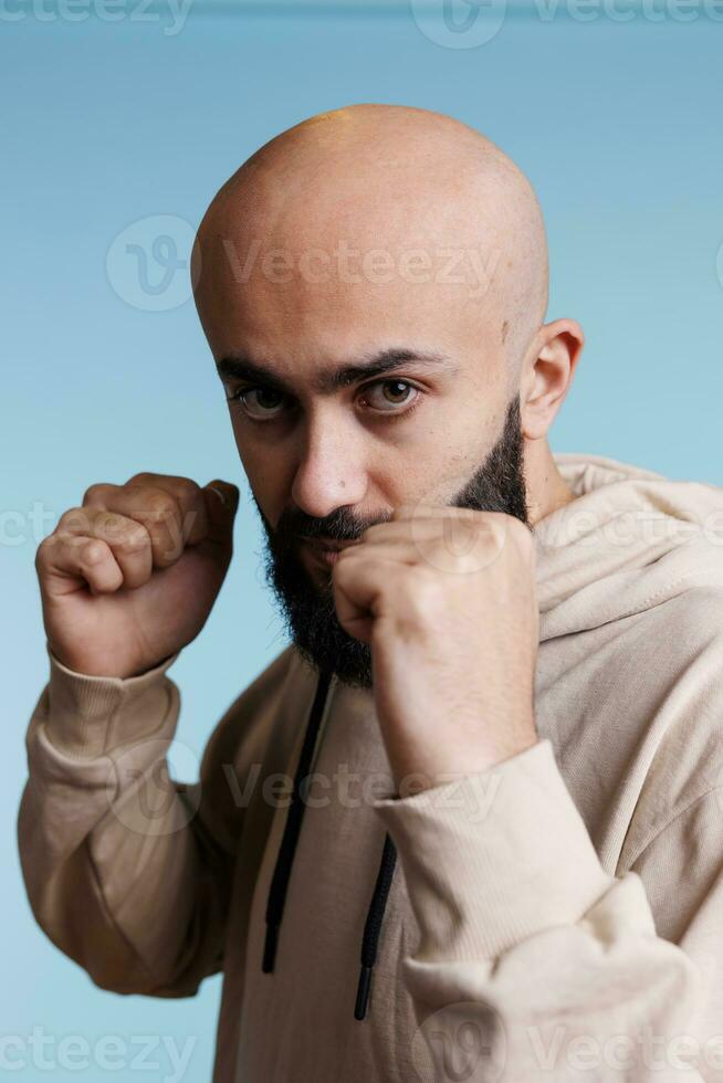 arabisch Mann zeigen Stärke während Stehen mit geballt Fäuste im Boxen Position Porträt. jung Person Herstellung Kämpfer Geste während suchen beim Kamera mit aggressiv Gesichts- Ausdruck foto