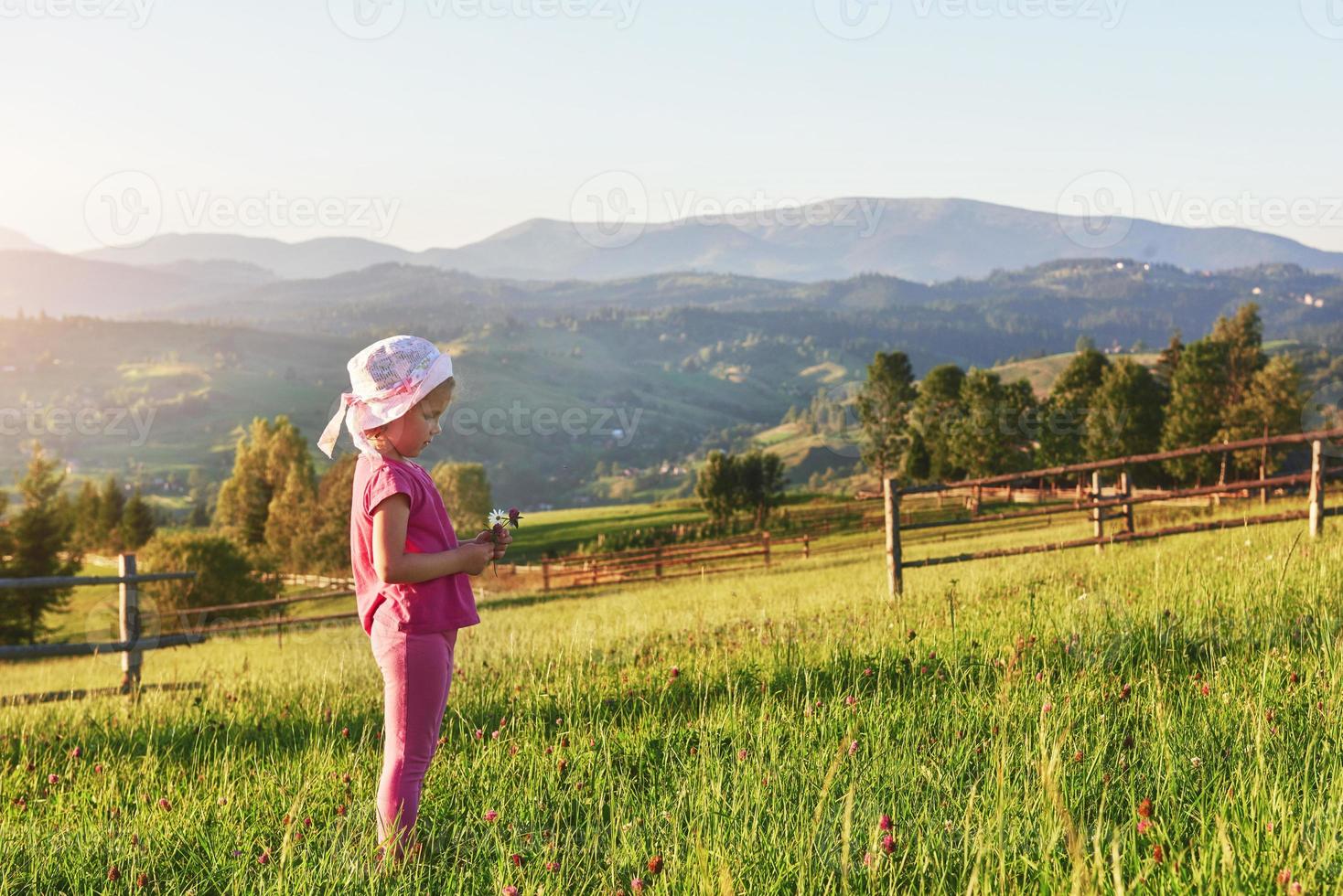 Süßes, glückliches kleines Mädchen, das draußen auf dem Rasen spielt und die Aussicht auf die Berge bewundern kann. Kopieren Sie Platz für Ihren Text foto