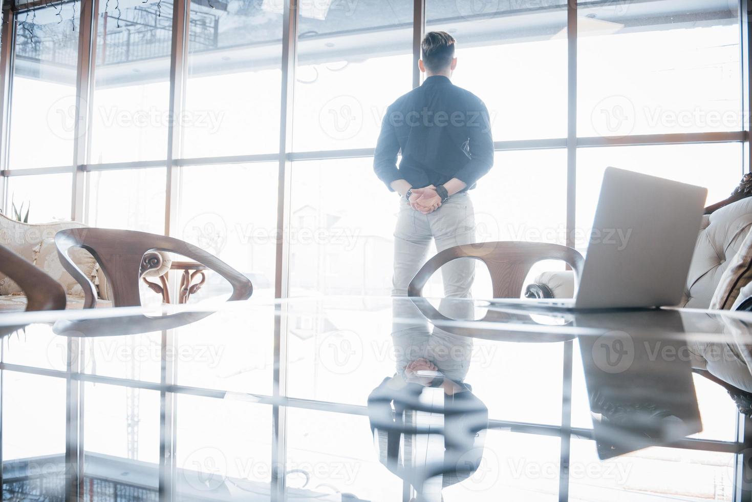 junger Geschäftsmann und Führer, der selbstbewusst im Büro im obersten Stockwerk steht und durch große Fenster auf die Stadt unten blickt foto