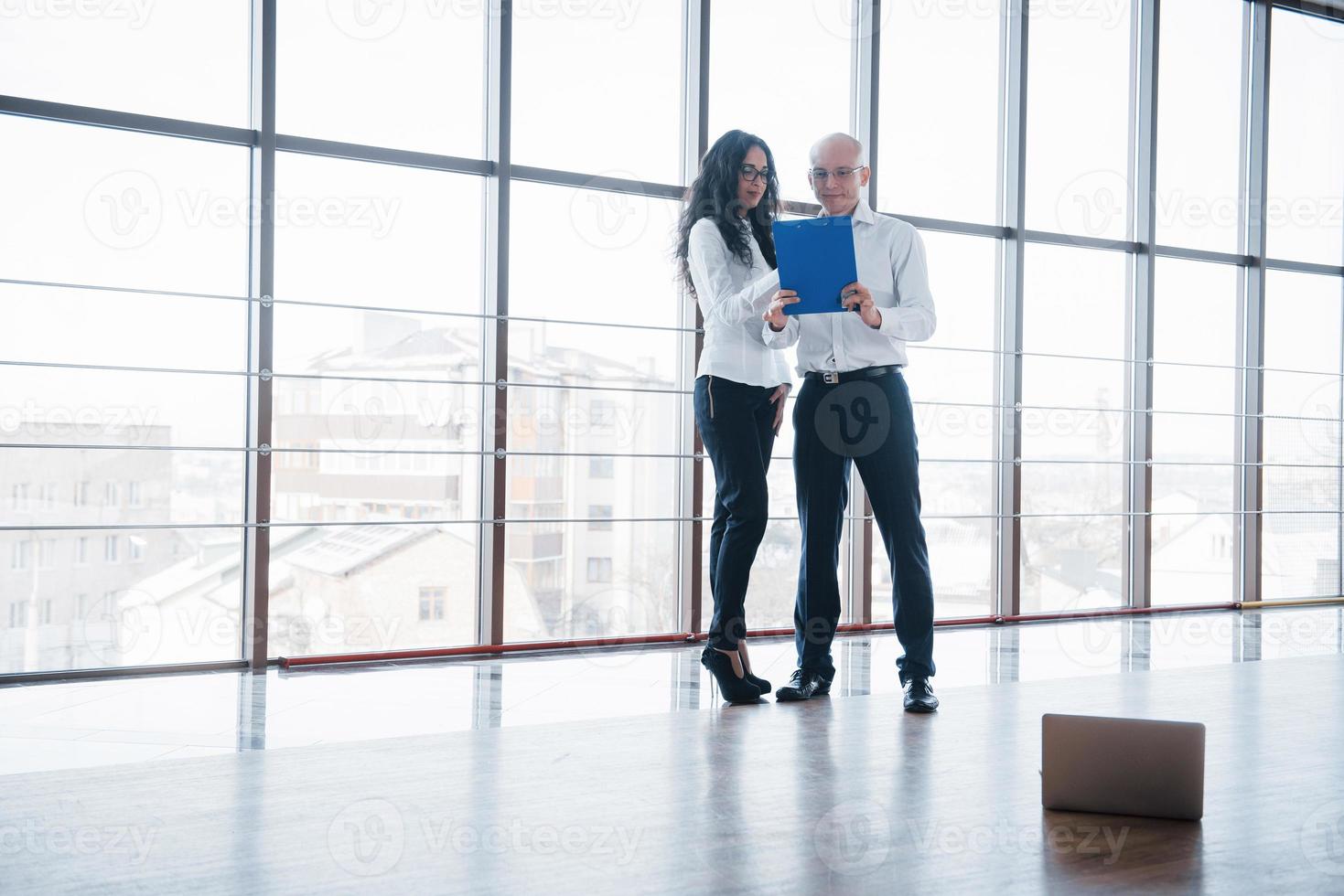 Geschäftsmann und Geschäftsfrau, die ein Diagramm auf den Platten- und Papierdokumenten am Fenster auf dem Hintergrund des Stadtbüros auf einem hohen Stockwerk studieren foto