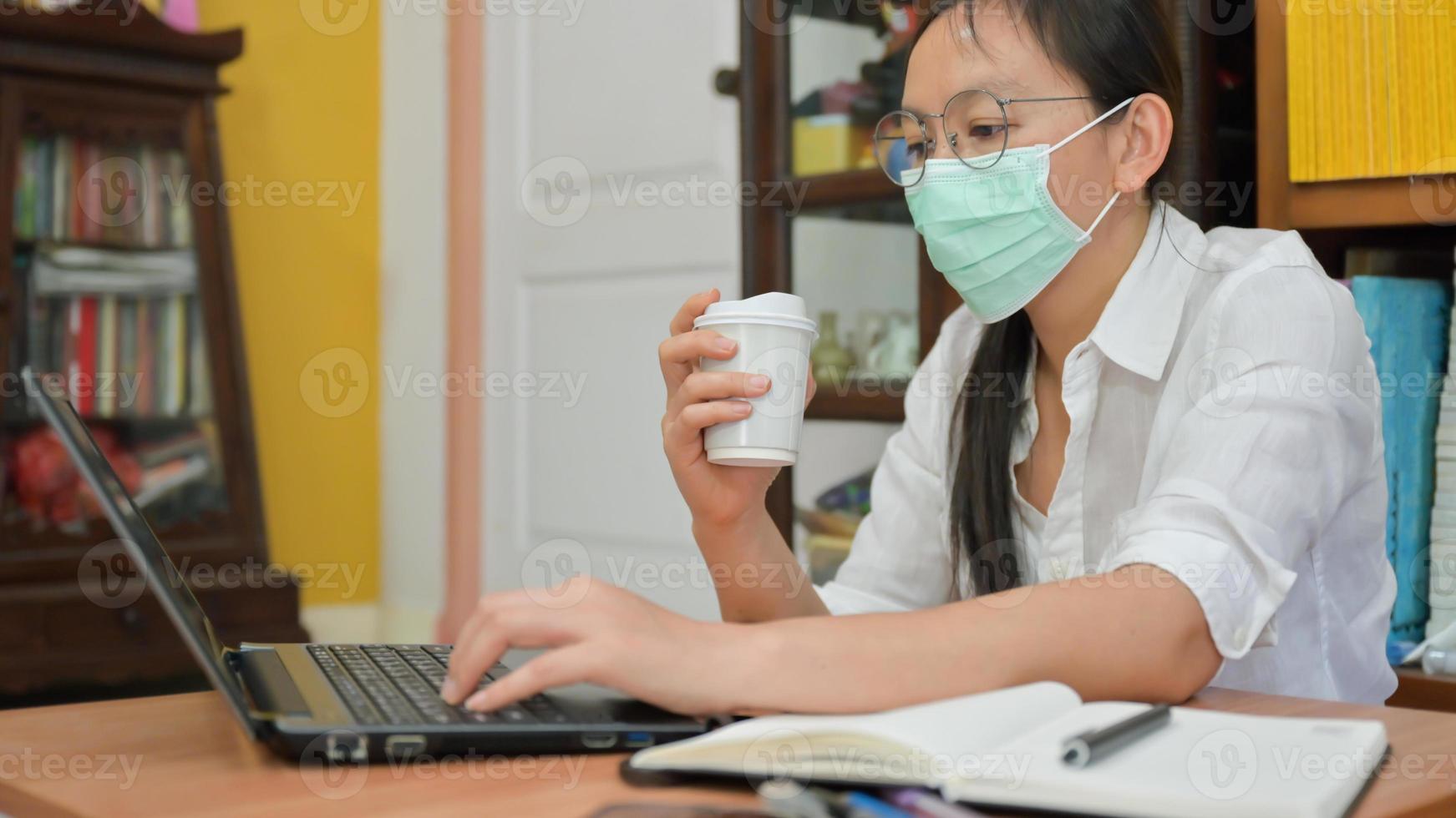 asiatische Frauenmaske, die eine Kaffeetasse hält und einen Laptop verwendet. Sie arbeitet zu Hause, um sich vor dem Koronavirus oder Covid-19 zu schützen. foto