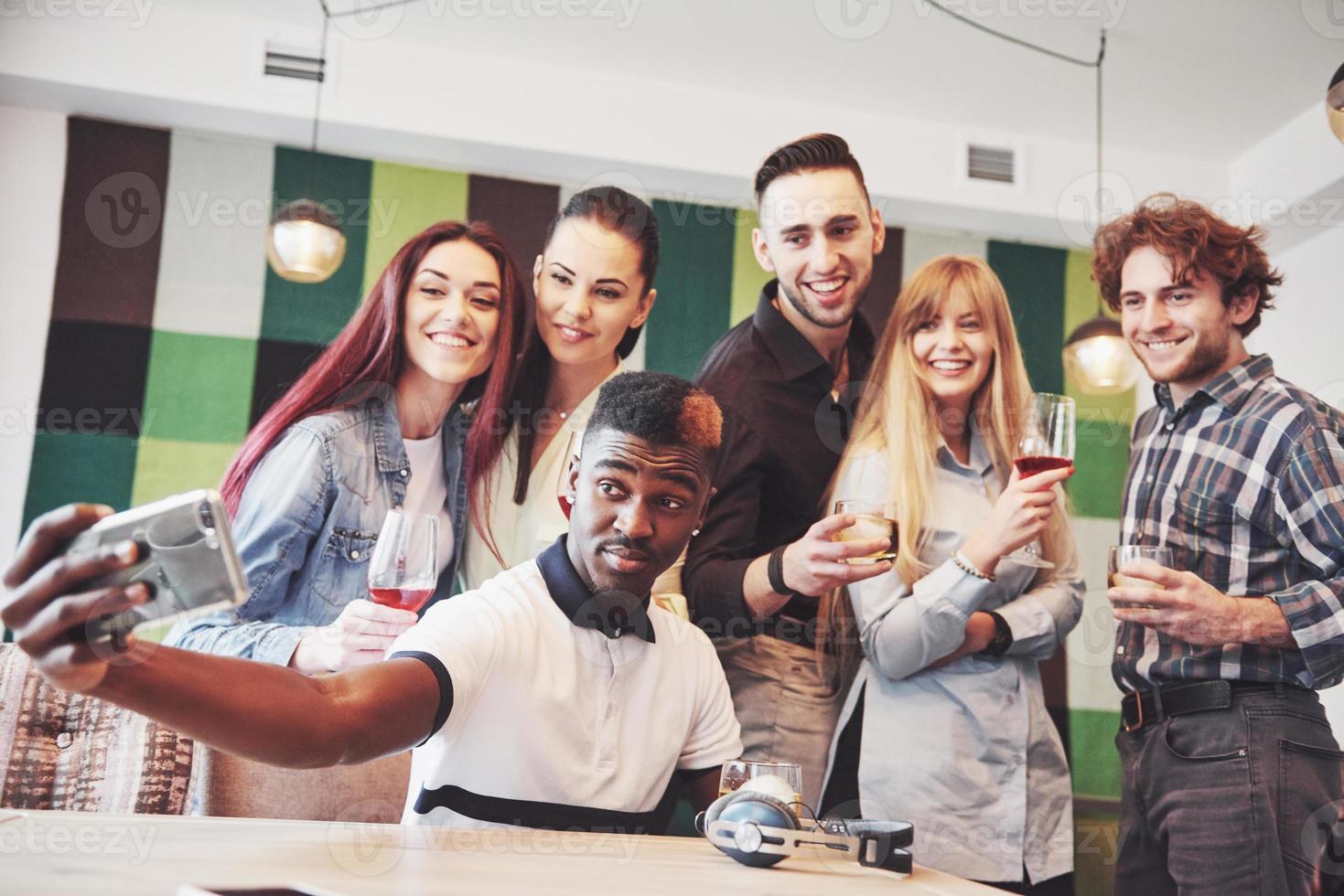 Freunde, die Spaß im Restaurant haben. Zwei Jungen und vier Mädchen, die trinken, Selfie machen, Friedenszeichen machen und lachen. im Vordergrund Frau mit Smartphone. alle tragen Freizeitkleidung foto