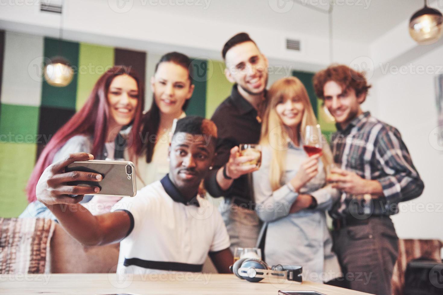 Freunde haben Spaß und machen Selfie im Restaurant. Zwei Jungen und vier Mädchen trinken Bier und essen Pizza mit frischem Salat zusammen. Junge mit Mütze macht Selfie mit seinem Handy. alle tragen Freizeitkleidung foto